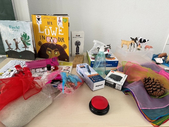 Auf diesem Bild sind verschiedene Materialien aus den Vorleseboxen für die inklusiven Vorleser:innen auf einem Tisch ausgelegt. Darunter sind Bücher, Tierfiguren, ein Buzzer für Unterstützte Kommunikation, Tücher, Sprühflasche, AnyBook Reader und viele weitere Dinge für das multisensorische Vorlesen.