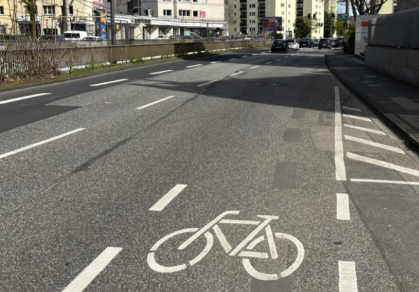 Eine dreispurige Straße mit einem Fahrrad-Symbol auf einer Spur, entfernt sind Autos und Häuser zu sehen.
